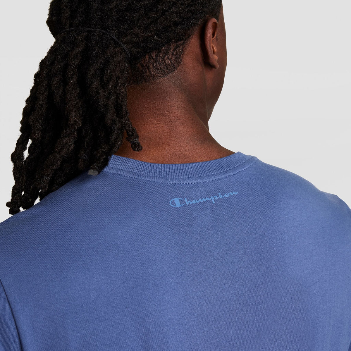 A person with long, dark dreadlocks, seen from behind, wears Champion’s Classic T-Shirt, C Logo in blue. The brand's logo appears near the neckline against a plain light background.