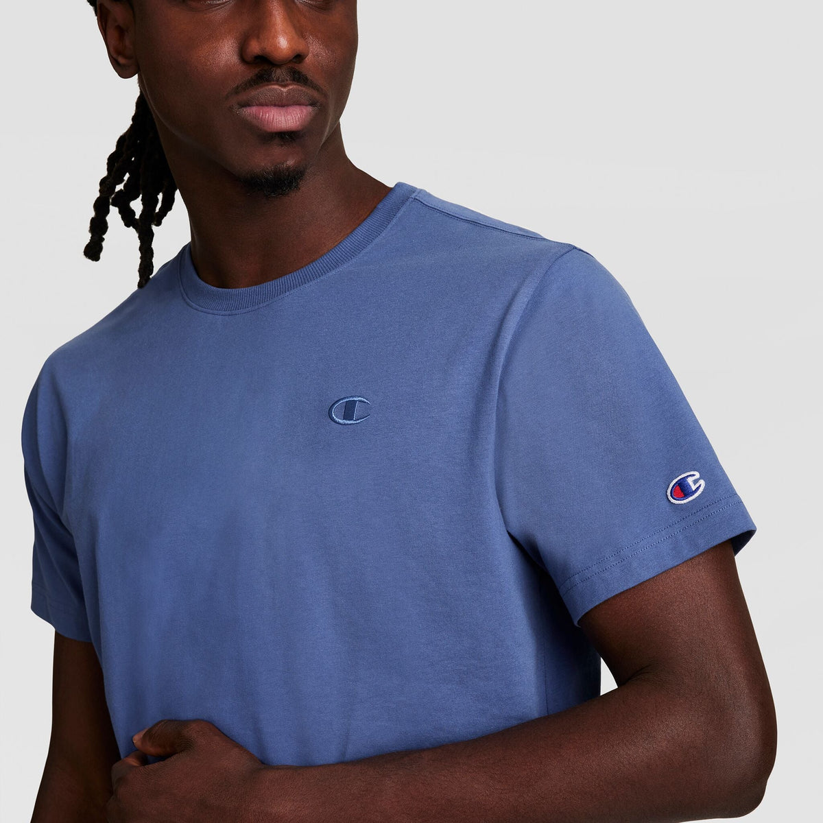 A man wears the Champion Classic T-Shirt, C Logo—blue cotton with the Champion logo on chest and sleeve—standing against a plain light gray background.