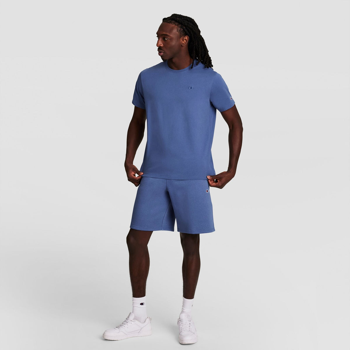 A man stands against a plain light gray background, wearing the Champion Classic T-Shirt with C Logo and matching blue shorts, paired with white socks and sneakers. He faces sideways with hands on his shorts, highlighting this iconic men's look.