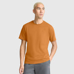 A man with light blond hair stands against a plain light background, wearing a Champion Classic T-Shirt, C Logo in orange paired with gray shorts. He has one hand in his pocket and gazes slightly to the side. The soft cotton fabric of the T-shirt ensures all-day comfort, adding to his effortlessly relaxed appearance.