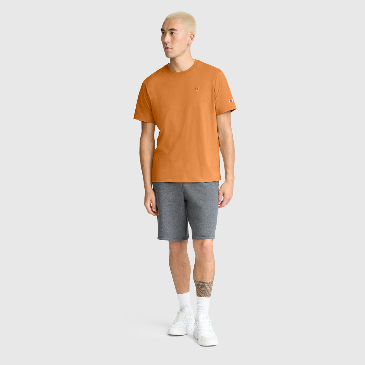 A person with short blond hair wears the Champion Classic T-Shirt, C Logo in orange, paired with gray shorts and white sneakers, standing against a plain gray background.