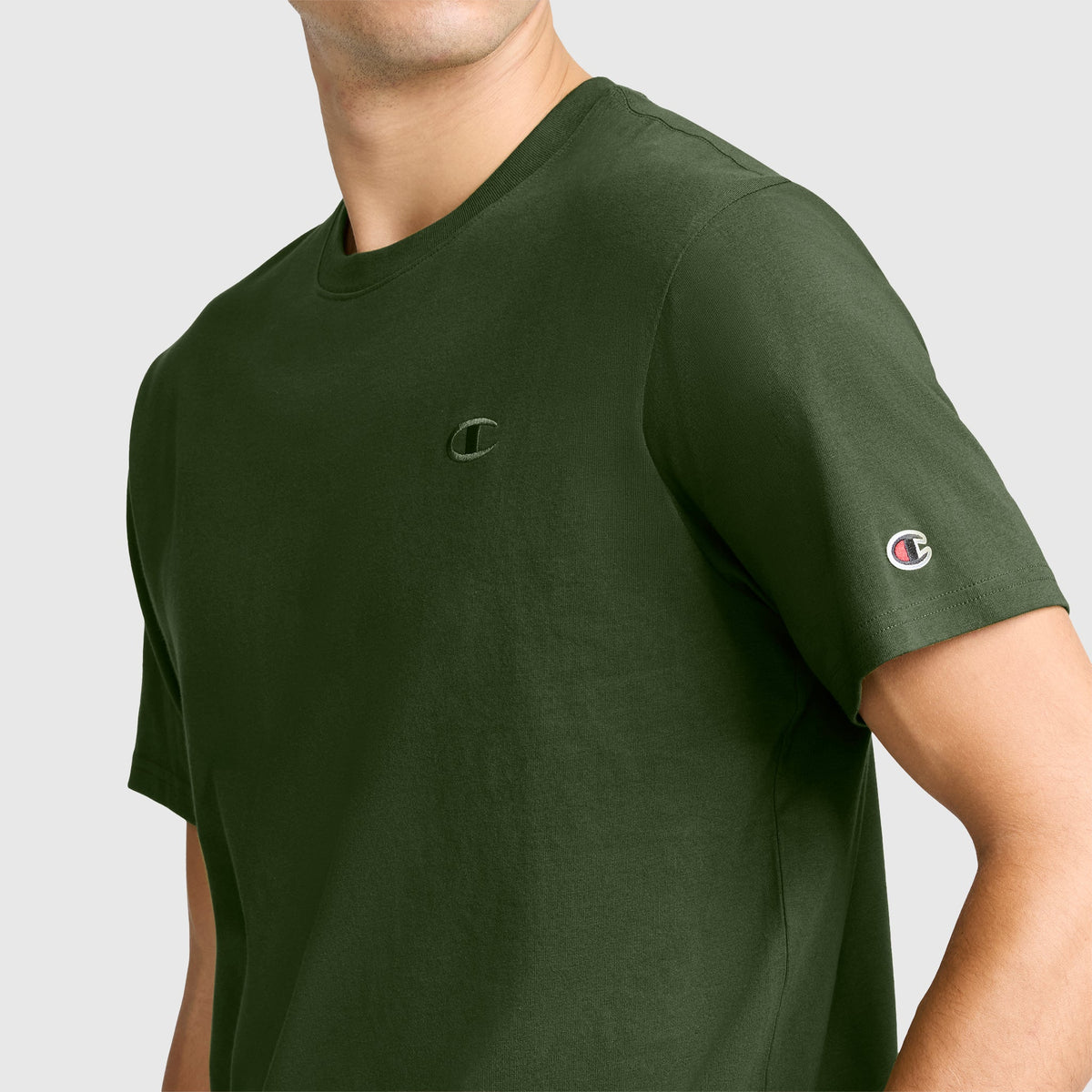 A man wears the Champion Classic T-Shirt, C Logo in dark green, featuring the iconic Champion branding on the chest and left sleeve. The shirt is set against a plain light gray background and is crafted from soft cotton for all-day comfort.