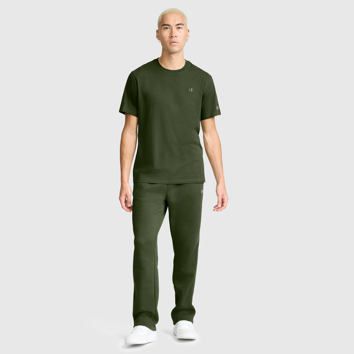 A person with short blond hair wears a dark green Champion Classic T-Shirt featuring the C Logo, paired with matching pants and white sneakers, standing against a plain light gray background.