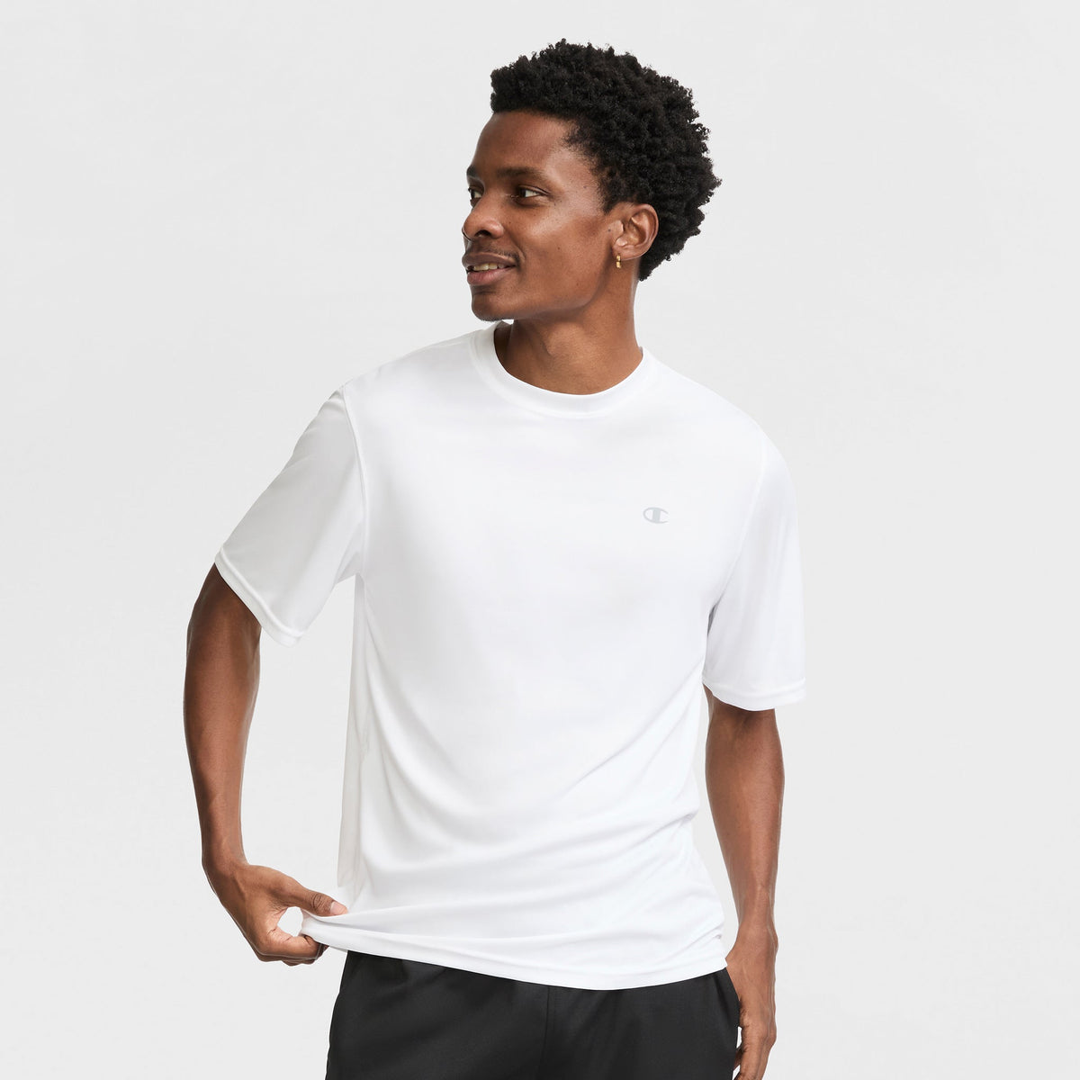 A man in a Champion Sport T-Shirt with Reflective C Logo and black shorts stands against a white background, holding his shirt's hem with one hand and smiling as he looks to his left.
