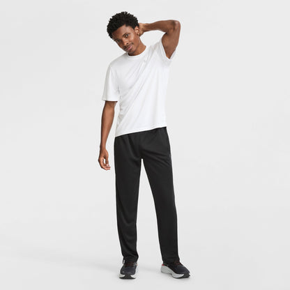 A man poses against a white background, smiling in a Champion Sport T-Shirt with a Reflective C Logo, paired with black pants and athletic shoes. He tilts his head and rests one hand on the back of his neck.