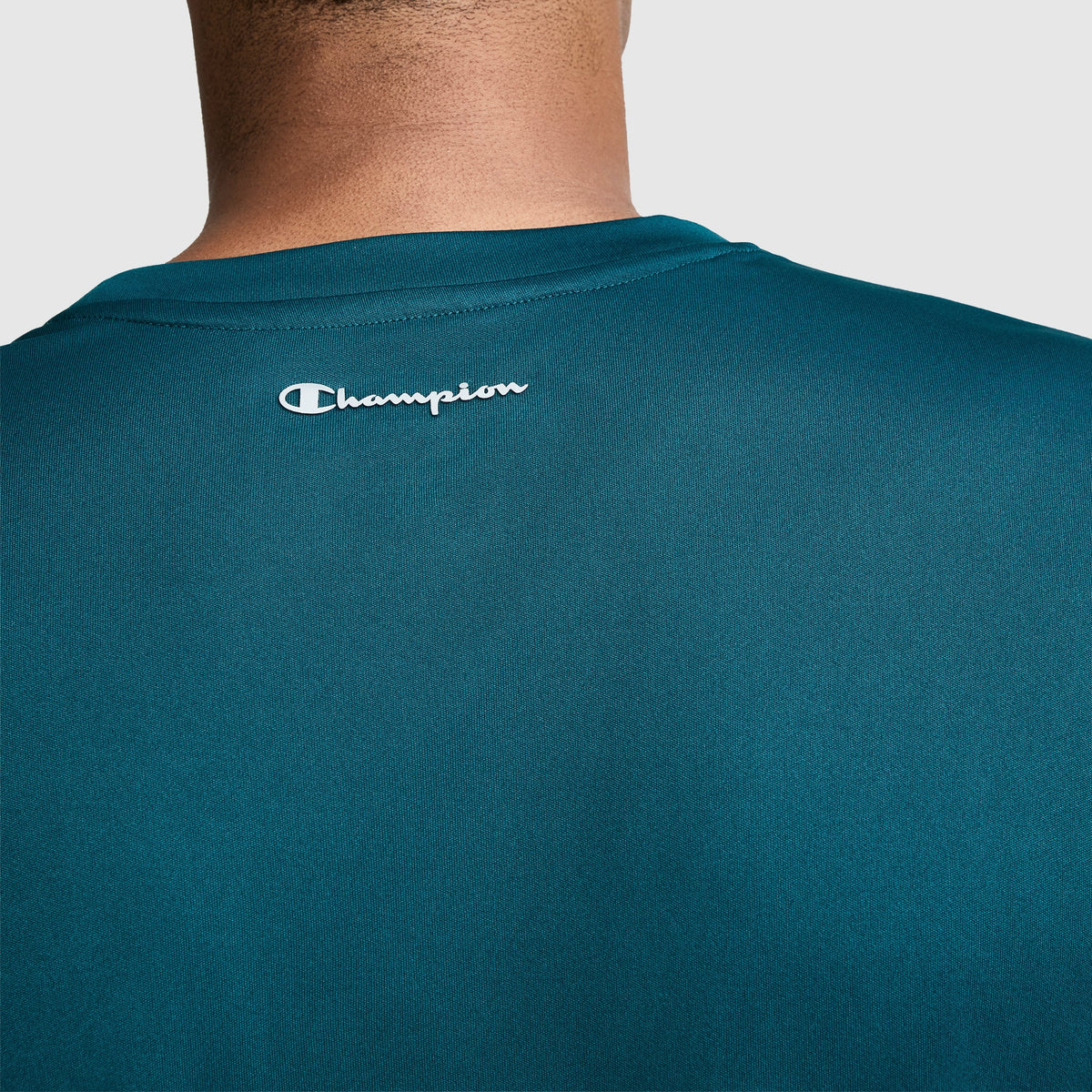 A close-up of a person wearing a teal Champion Sport T-Shirt, Reflective C Logo, with the white Champion logo displayed on the upper back. The image highlights the back of this men’s athletic shirt and the wearer’s neck.