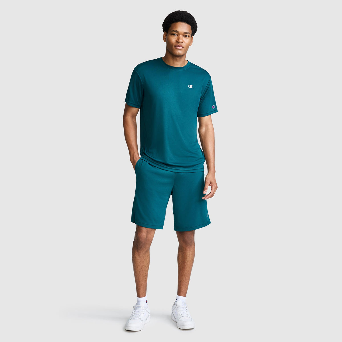 A man stands against a light gray background wearing the Champion Sport T-Shirt with Reflective C Logo in teal, paired with matching athletic shorts and white sneakers. The Sport T-Shirt is designed with moisture-wicking fabric for all-day comfort. He has one hand in his pocket and is smiling slightly.