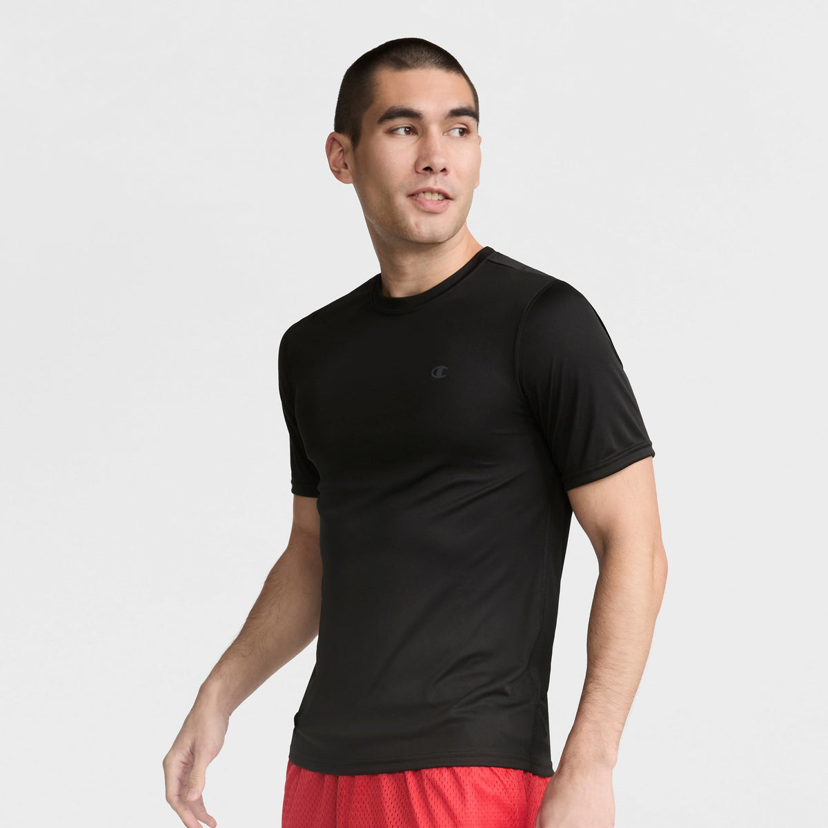 A man with short dark hair wears a fitted black Champion Sport T-Shirt with Reflective C Logo and red shorts, standing against a plain light background and looking to his left with a neutral expression.