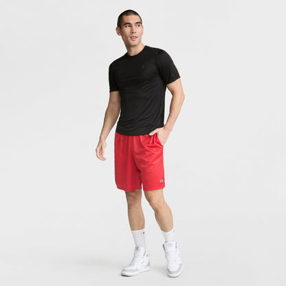A man stands against a white background wearing a Champion Sport T-Shirt with Reflective C Logo, red athletic shorts, white crew socks, and white sneakers. He has one hand in his pocket and looks slightly to the side.