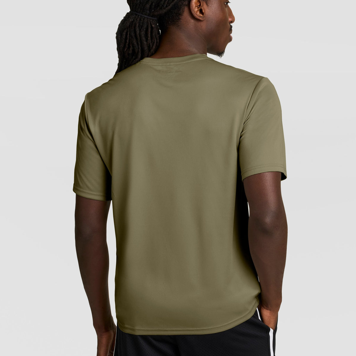A man with long braided hair, seen from behind, wears a Champion Sport T-Shirt with Double Dry technology and a reflective C logo in olive green, paired with black shorts, standing against a plain light gray background.