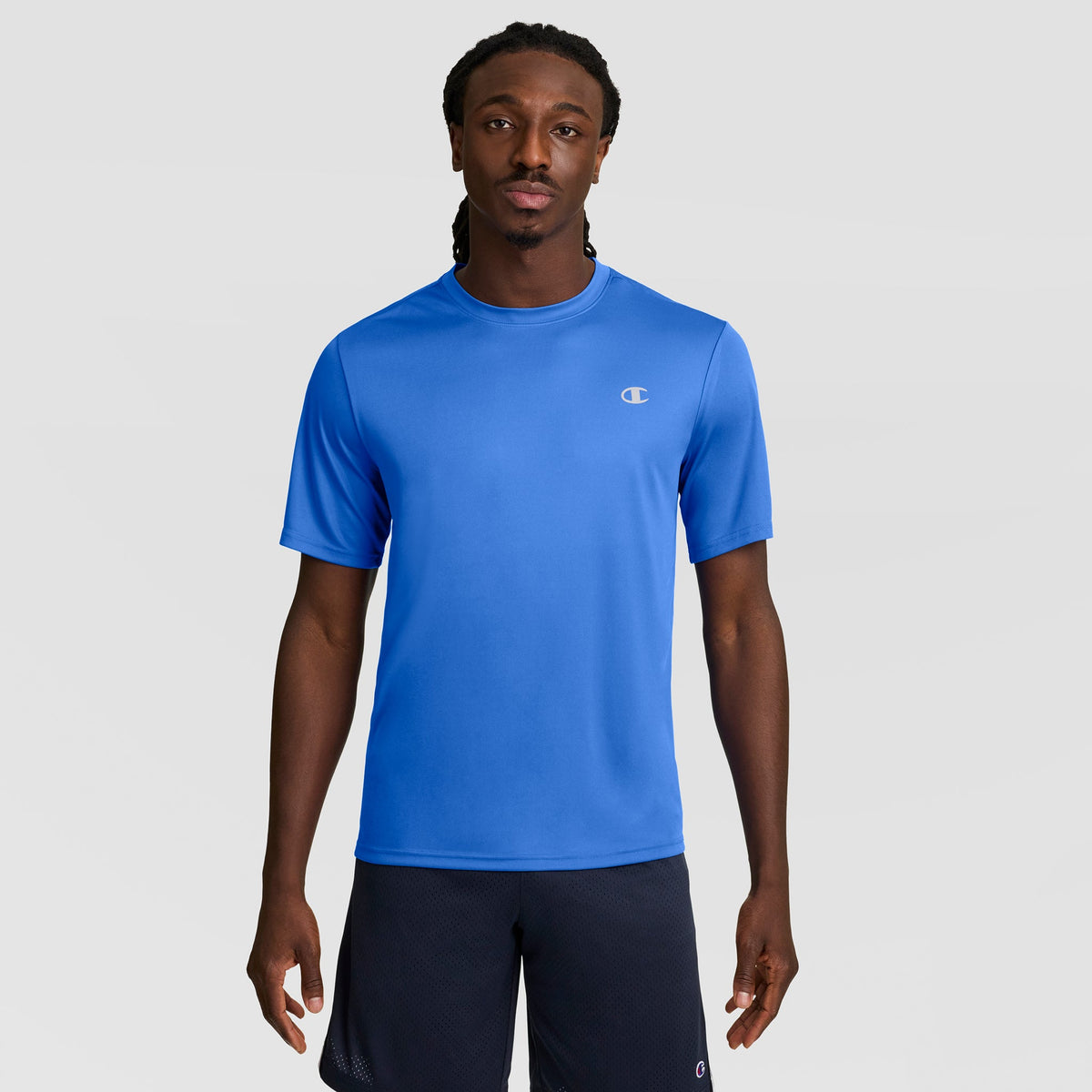 A man with long braided hair models a Champion Sport T-Shirt with Reflective C Logo in bright blue and black shorts, standing against a plain light gray background, facing forward with a neutral expression for an athletic look.