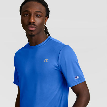 A man with long braided hair wears a bright blue Champion Sport T-Shirt featuring a reflective C logo, standing against a plain light gray background and looking at the camera.