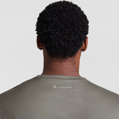 A person with short curly hair is shown from behind in a Champion Sport T-Shirt, Reflective C Logo - Big & Tall, olive green, featuring the logo on the upper back. The background is plain and light-colored.