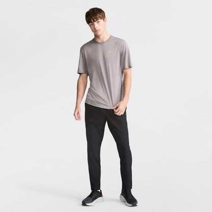 A young man with straight brown hair and a neutral expression stands against a white background, wearing a loose-fitting gray Champion Sport T-Shirt with a reflective C logo, black athletic pants, and black sneakers with white soles.