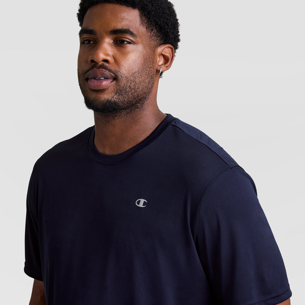 A man models the Champion Sport T-Shirt, Reflective C Logo - Big & Tall in dark navy against a light background. The moisture-wicking crewneck features the reflective C logo on the left chest.