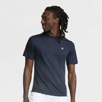 A man with long, dark dreadlocks wears a Champion Sport T-Shirt with Reflective C Logo in navy blue and white shorts, looking to his left against a plain light gray background.