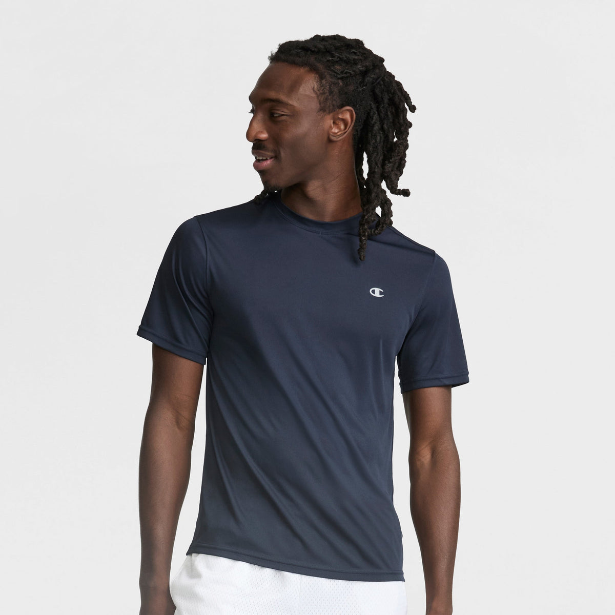 A man with long, dark dreadlocks wears a Champion Sport T-Shirt with Reflective C Logo in navy blue and white shorts, looking to his left against a plain light gray background.
