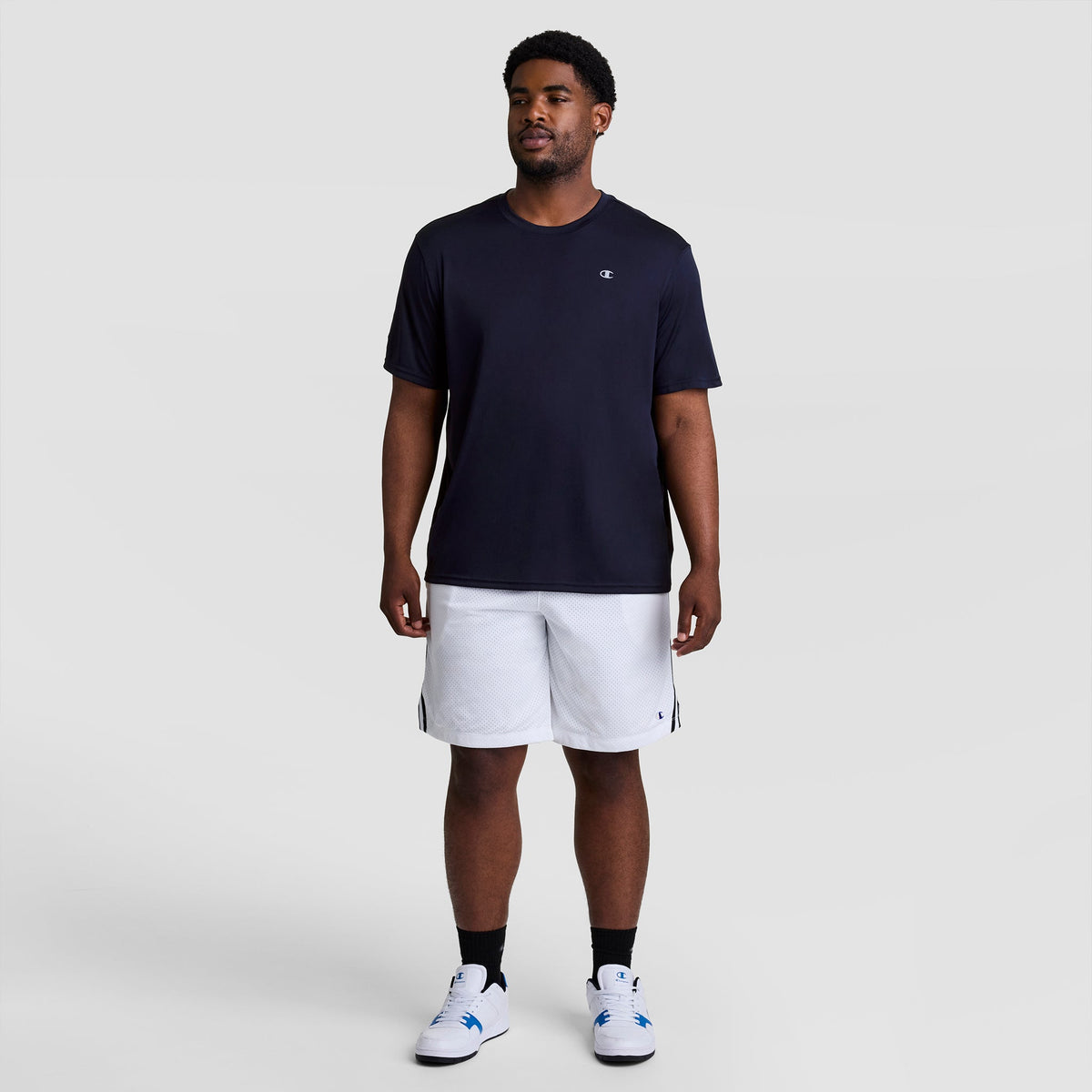 A person wearing a dark blue Champion Sport T-Shirt, Reflective C Logo - Big & Tall with a small logo on the chest and white athletic shorts.