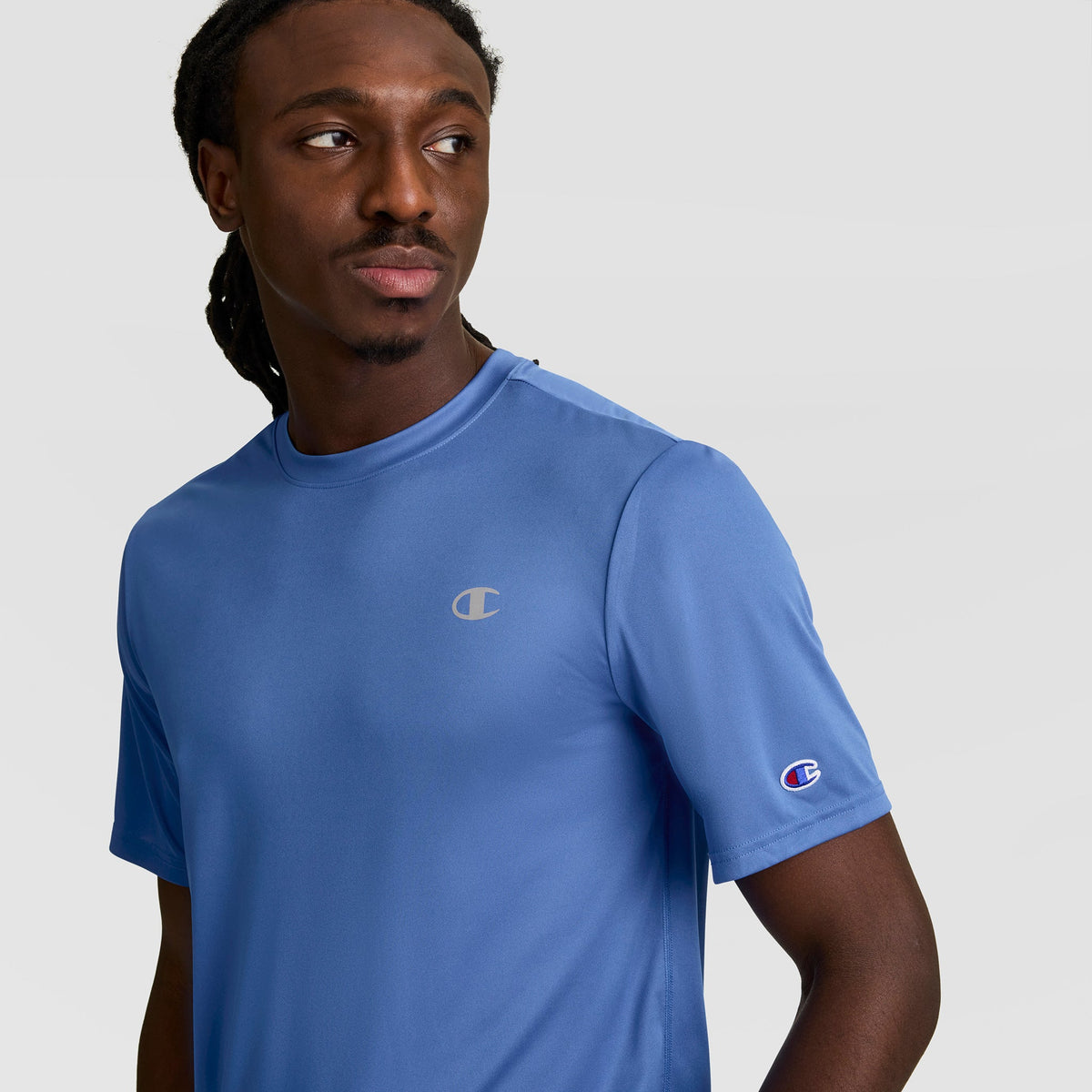 A man with long dark hair wears a Champion Sport T-Shirt, Reflective C Logo, in light blue. The short-sleeve tee features logos on the chest and sleeve and offers comfort with Double Dry technology. He looks left against a plain backdrop.