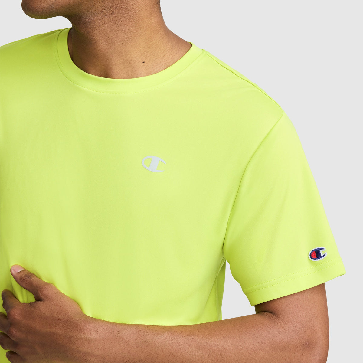 A person wearing a bright yellow-green Champion Sport T-Shirt, Reflective C Logo, featuring the Champion logo on the chest and sleeve, is seen from neck to waist with their left hand resting on their torso.