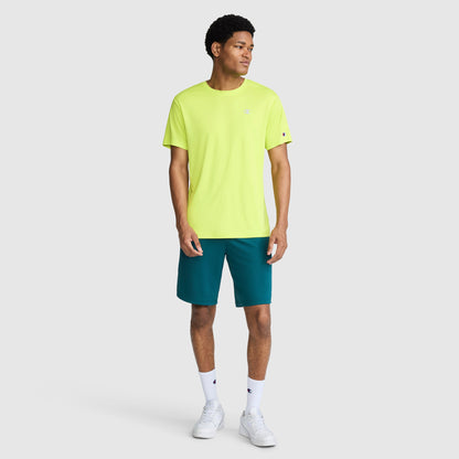 A man stands against a plain light gray background, wearing a bright yellow Champion Sport T-Shirt with a Reflective C Logo, teal shorts, white socks, and white sneakers. He is looking to his left with his arms relaxed by his sides.