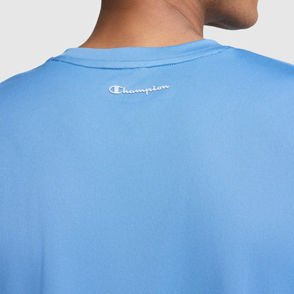 Close-up of a person wearing a light blue Champion Sport T-Shirt, Reflective C Logo, showing the back of the shirt with the white reflective Champion logo near the neckline. The person’s head and neck are partially visible.