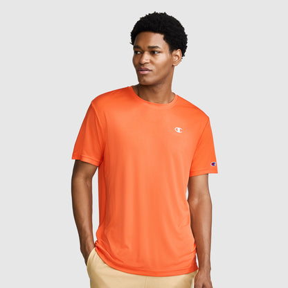 A man wearing a Champion Sport T-Shirt with a Reflective C Logo in orange and beige pants stands against a plain gray background, looking slightly to his left with one hand in his pocket.