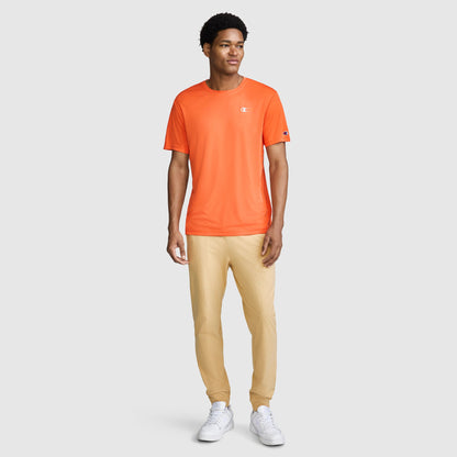 A person stands facing forward, wearing a Champion Sport T-Shirt, Reflective C Logo in orange—featuring moisture-wicking fabric and anti-odor protection—paired with beige pants and white sneakers against a plain light-colored background.