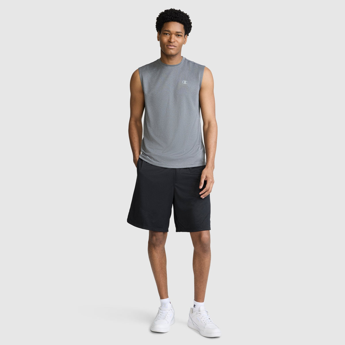 A man stands against a plain background wearing the Champion Sport Muscle Tank with a Reflective C Logo, paired with black shorts and white sneakers. This moisture-wicking sleeveless workout shirt keeps him comfortable as he looks at the camera with his hands by his sides.