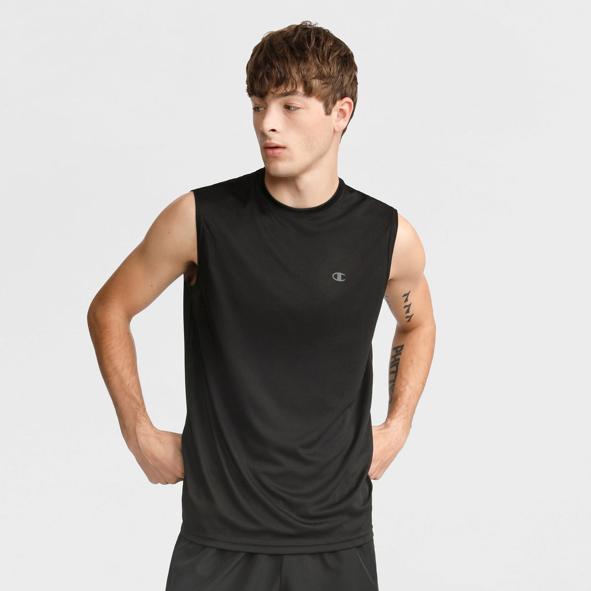 A young man stands hands on hips, looking to the side against a plain light background, wearing the Champion Sport Muscle Tank with Reflective C Logo and black shorts.