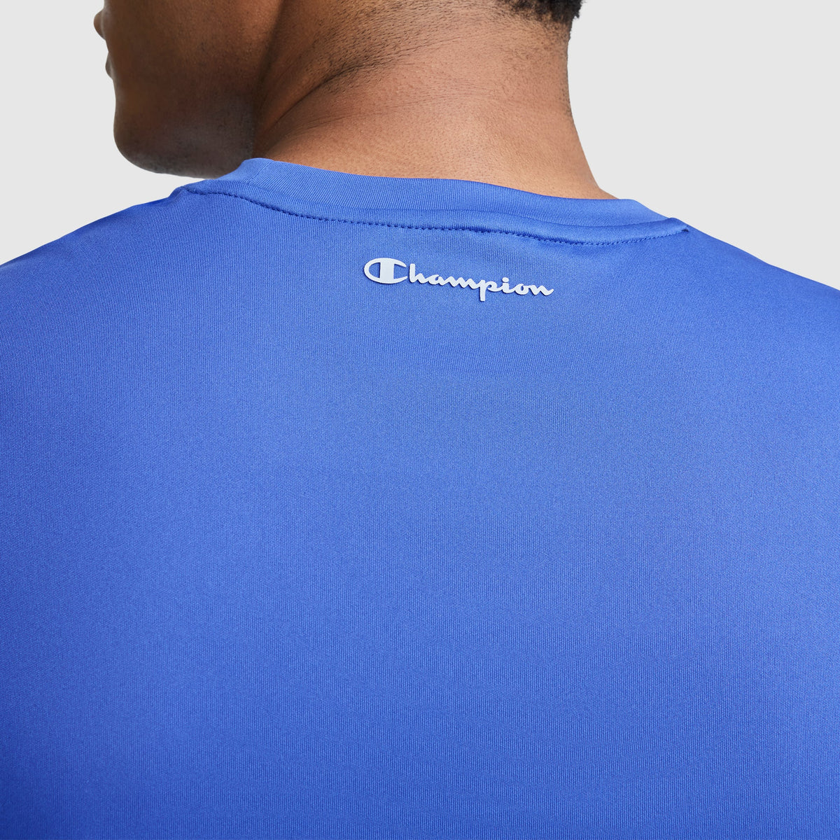 A close-up of the back of a person wearing a blue Champion Sport Muscle Tank, Reflective C Logo. This sleeveless men's workout shirt features the white reflective Champion logo near the collar and is crafted with moisture-wicking fabric to help keep you cool and comfortable during your workout.