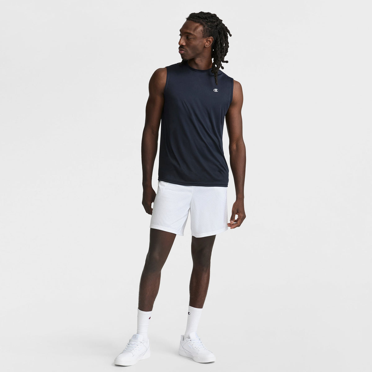 A man stands against a plain white background wearing the Champion Sport Muscle Tank with Reflective C Logo, paired with white shorts, socks, and sneakers. He looks to the side with one hand relaxed and the other by his side.