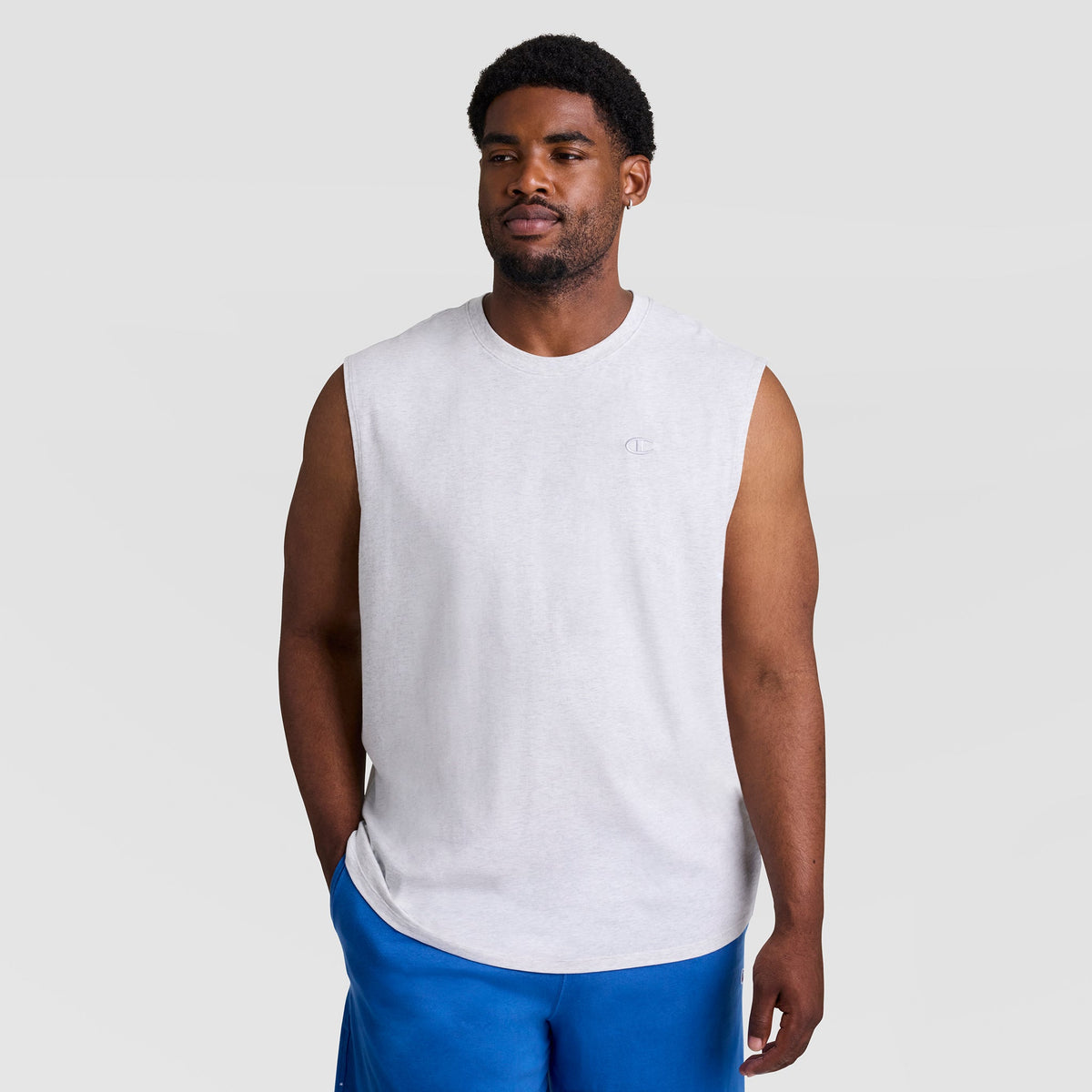 A man wears the Champion Muscle Tank, C Logo - Big & Tall, paired with blue shorts and stands against a plain light gray background, looking slightly to the side with a relaxed expression.