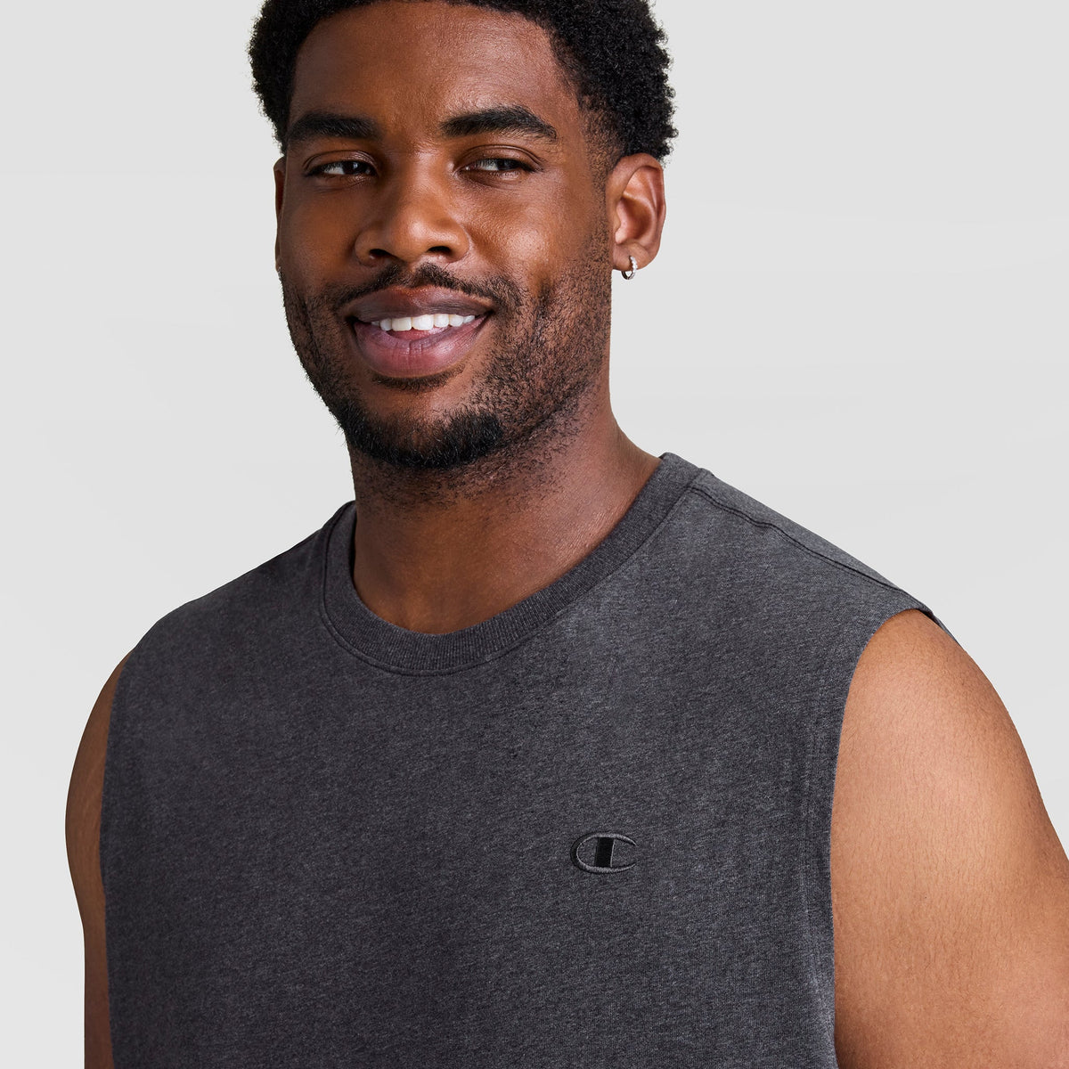 A man with short curly hair, a trimmed beard, and a small hoop earring smiles while wearing the Champion Muscle Tank, C Logo - Big & Tall in dark gray against a plain light gray background.