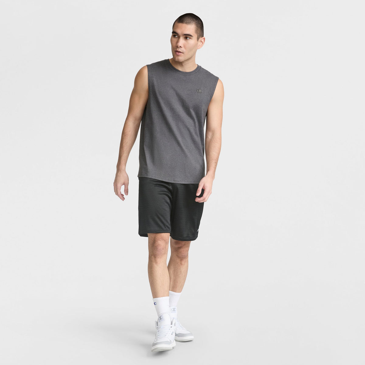 A man wearing a gray Champion Muscle Tank, black athletic shorts, white crew socks, and white sneakers stands looking left against a plain white background.