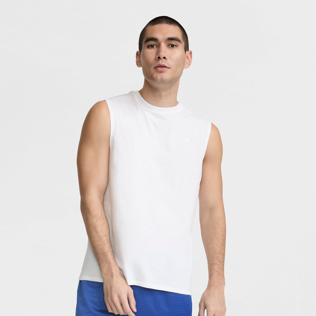 A man with short dark hair wears a Champion Muscle Tank, C Logo, and blue athletic shorts, standing against a plain light background and looking slightly to the side.