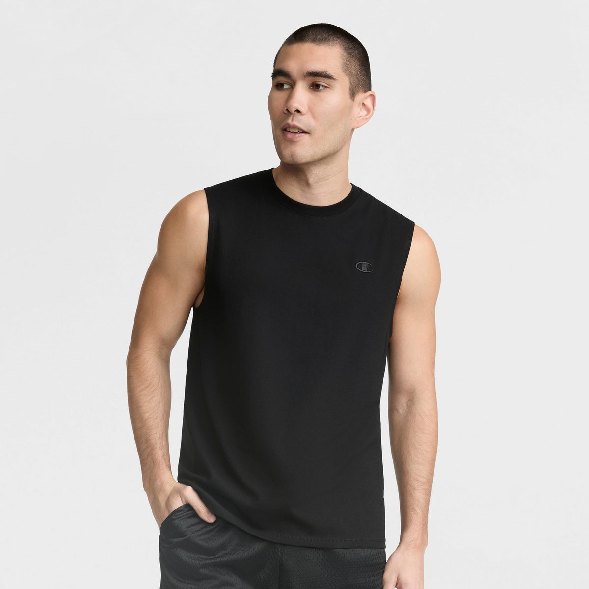 A man with short dark hair wears a black Champion Muscle Tank (C Logo) and black shorts, standing with one hand in his pocket against a plain white background.