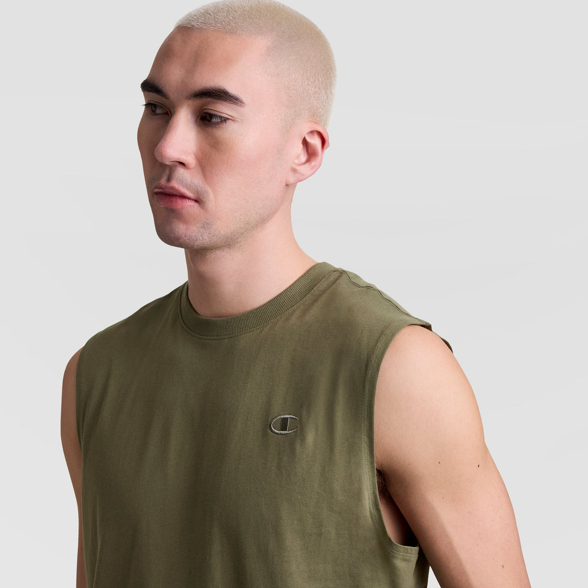A person with short, bleached blond hair models the Champion Muscle Tank, C Logo—an olive green muscle tee with a small embroidered chest logo—against a plain light gray backdrop.