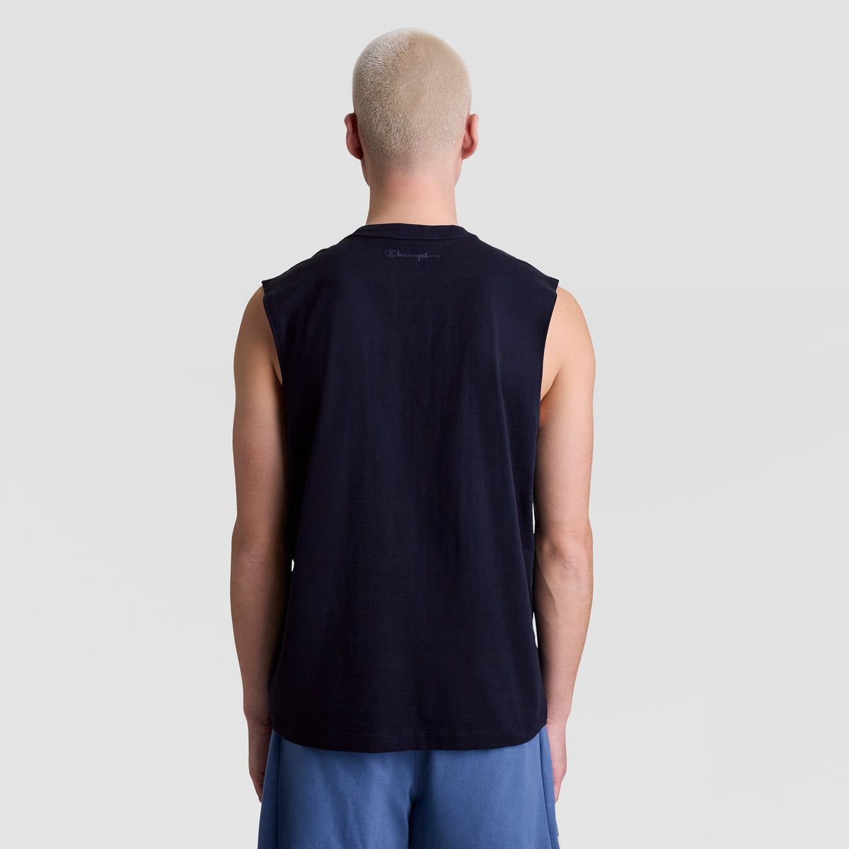 A person with short blond hair is shown from behind, wearing a black Champion Muscle Tank (C Logo) and blue shorts, standing against a plain light gray background.