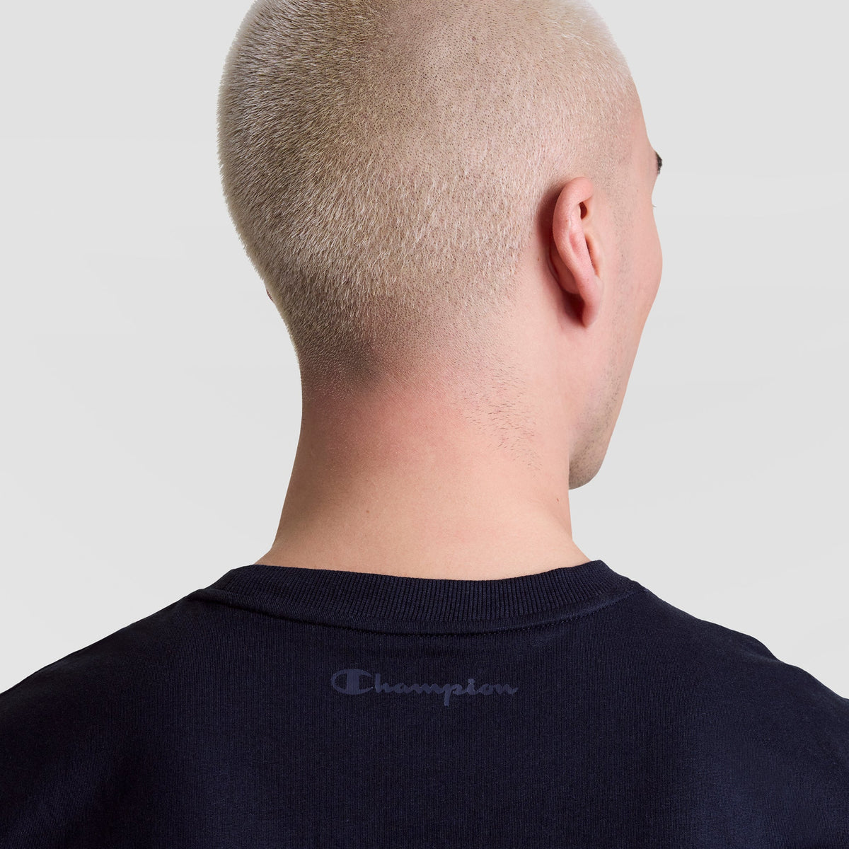 A person with very short, platinum blonde hair is shown from behind, wearing a dark navy Champion Muscle Tank, C Logo. "Champion" is printed on the upper back. The background is plain and light gray.