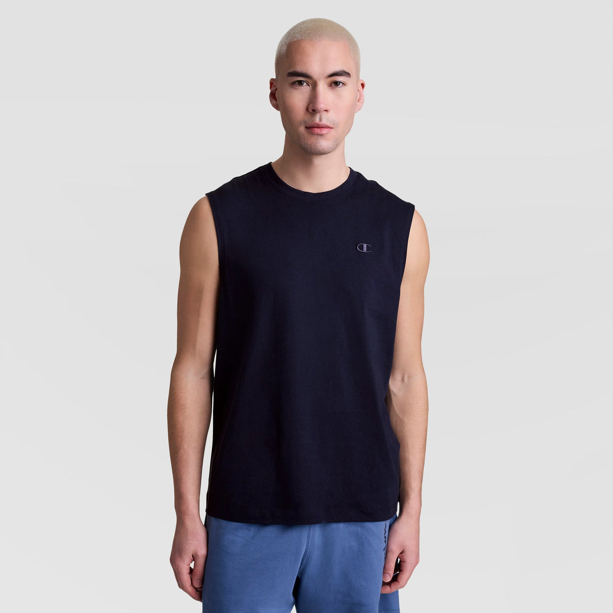 A person with short, light blond hair models the Champion Muscle Tank, C Logo—a sleeveless black top—paired with blue shorts against a plain light gray background, looking straight at the camera with a neutral expression.