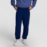 A person stands hands-on-hips in Champion’s Modern Retro Washed Pant with C Logo and a blue Champion sweatshirt. White sneakers finish the look. The face is not visible, and the background is plain and light.
