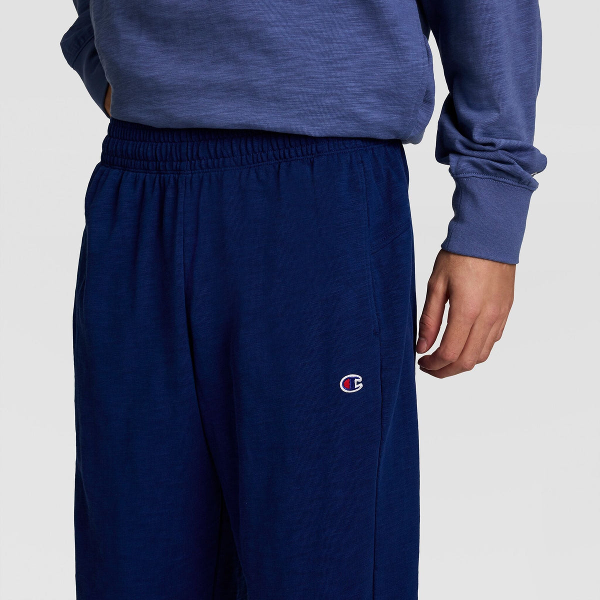A person in navy blue Champion Modern Retro Washed Pant, C Logo, their right hand relaxed at their side. Only the lower torso and upper legs are visible.