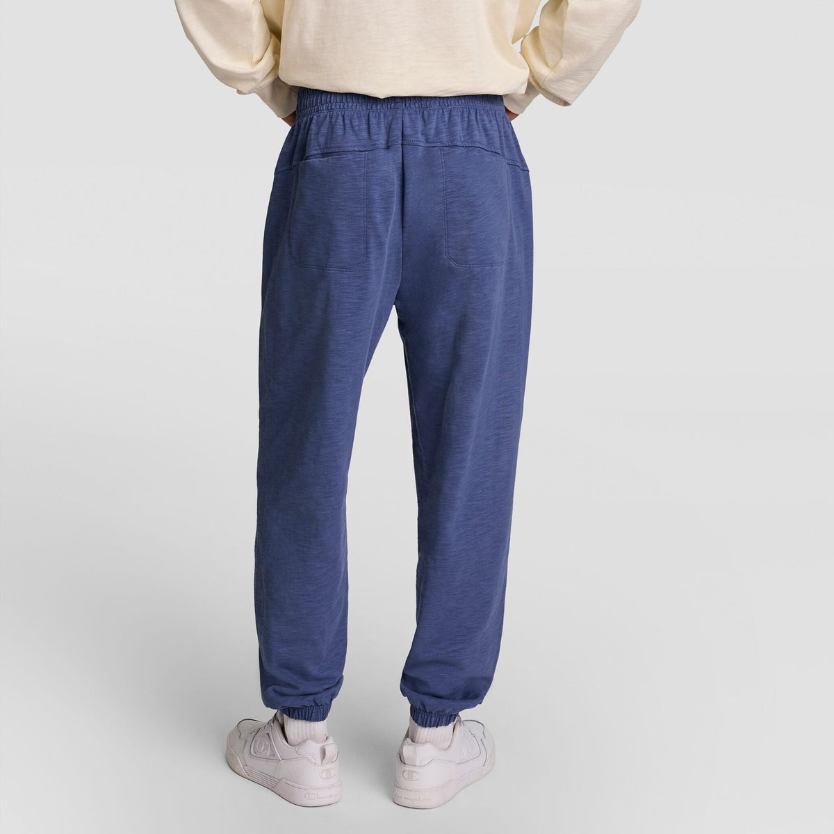 A person is shown from the back wearing a cream sweatshirt, Champion’s Modern Retro Washed Pant, C Logo in blue, and white sneakers, standing on a plain light background.