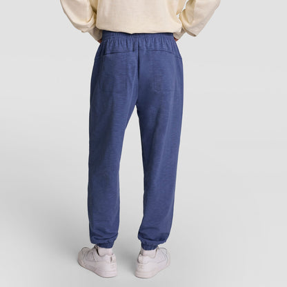 A person is shown from the back wearing a cream sweatshirt, Champion’s Modern Retro Washed Pant, C Logo in blue, and white sneakers, standing on a plain light background.