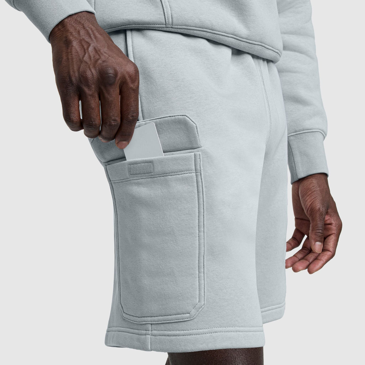 A person in light gray Champion 8" Fleece Cargo Shorts with a C Logo and a matching sweatshirt places their smartphone into the shorts' rectangular side pocket.