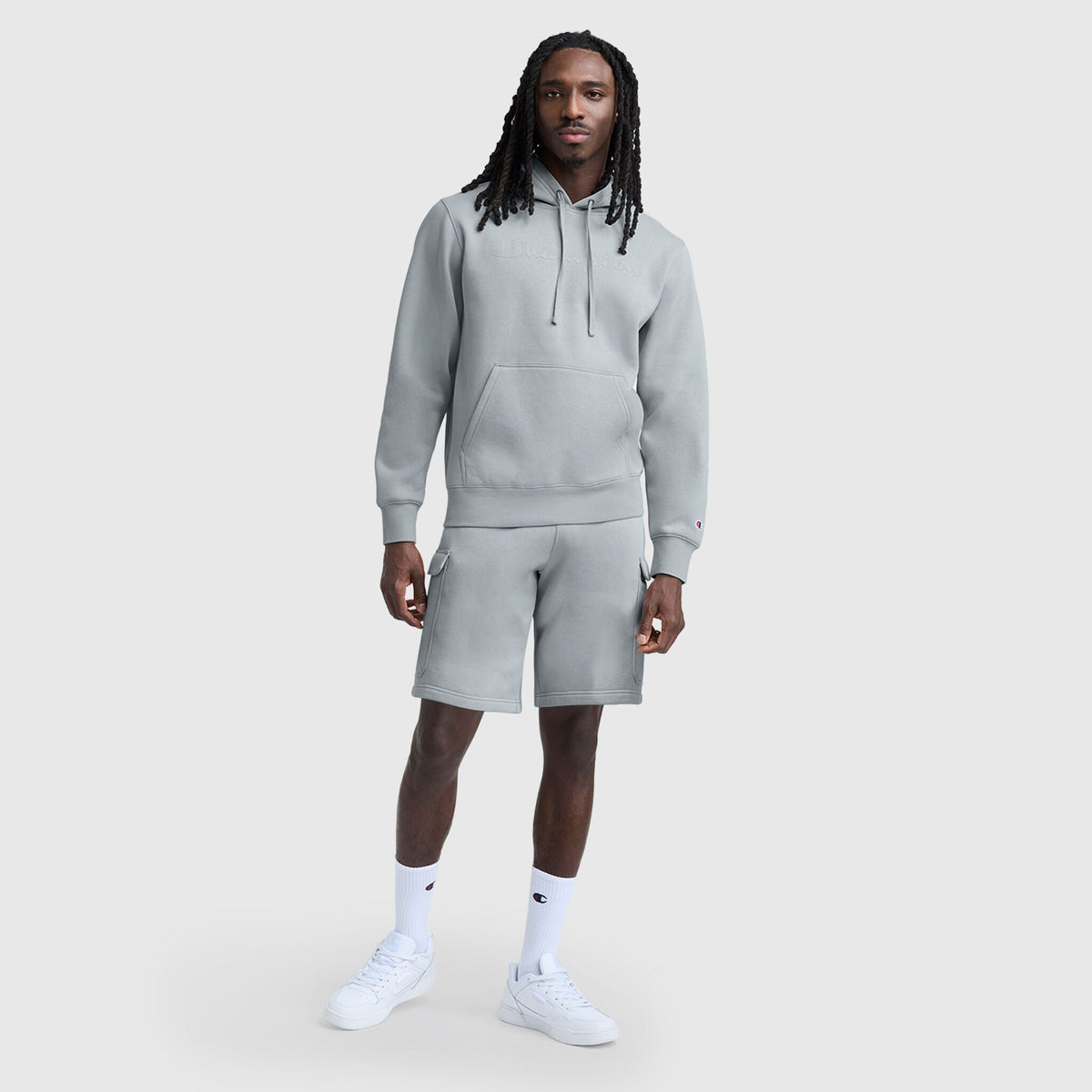 A man with long dreadlocks stands against a plain light gray background wearing a light gray hoodie, Champion 8" Fleece Cargo Short with C Logo, white socks, and white sneakers.