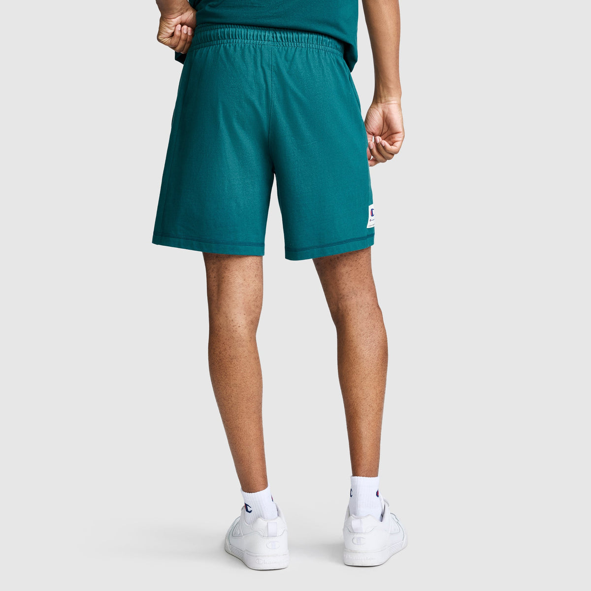 A person is shown from the back wearing Champion 7" Jersey Shorts with the iconic script logo, paired with a matching teal shirt, white socks also featuring the Champion script logo, and white sneakers. The background is plain and light-colored.