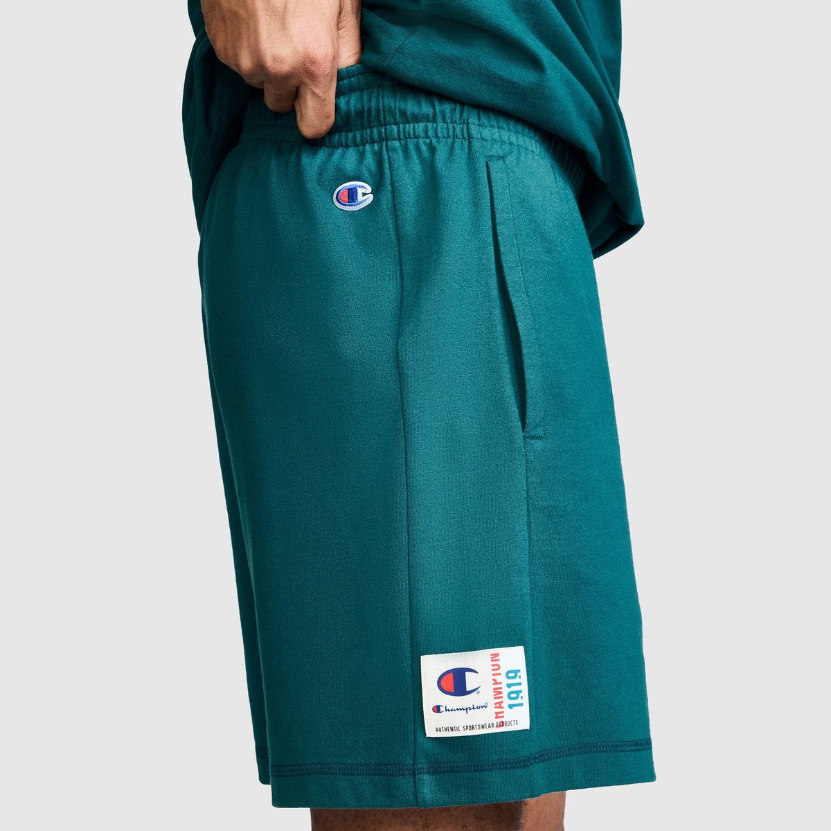 A person is wearing Champion's 7" Jersey Shorts with a script logo in teal, made from soft heritage cotton. The shorts feature a logo patch near the hem and a small Champion script logo by the waistband. Their hand rests casually on the waistband, set against a plain gray background.