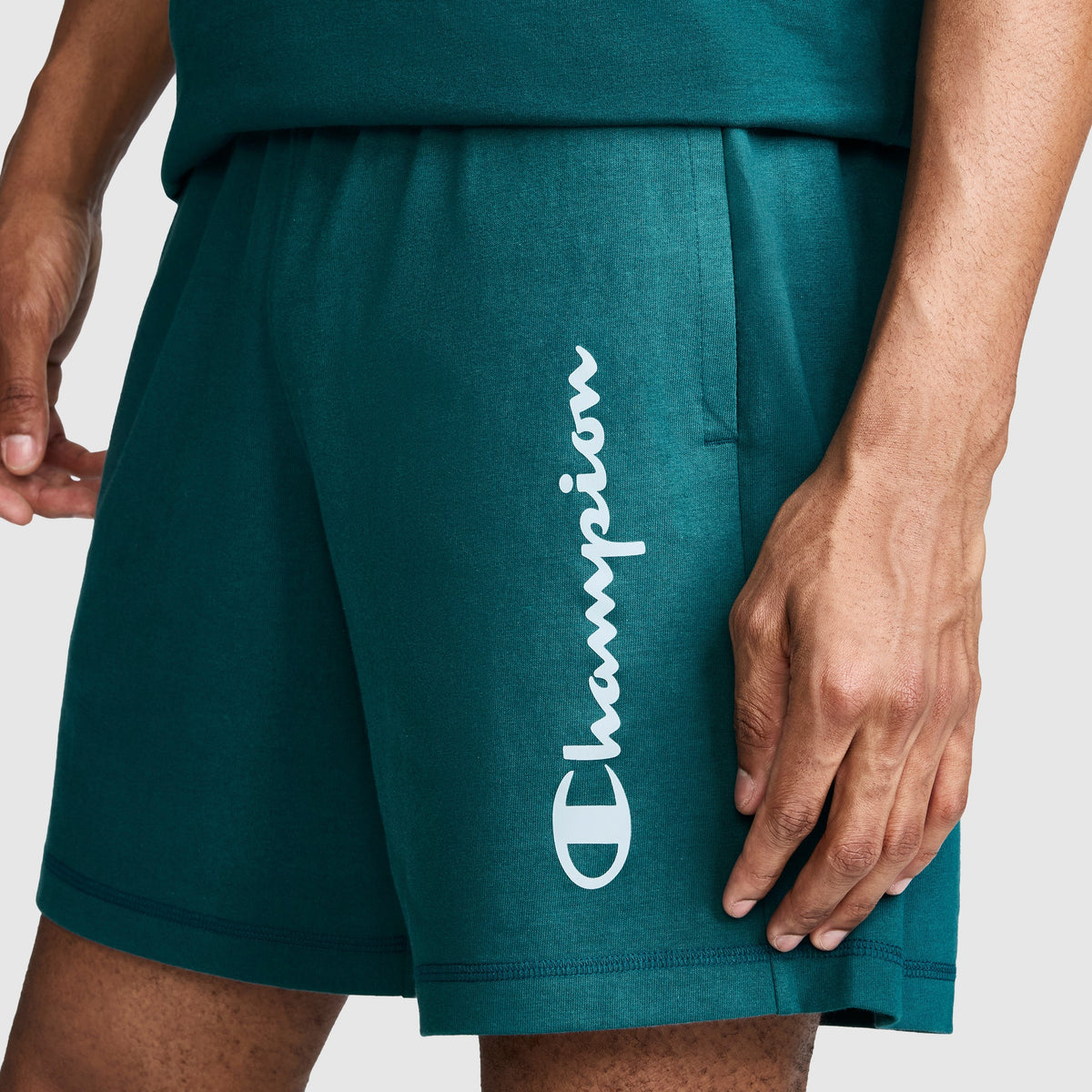 Wearing Champion’s 7" Jersey Shorts, Script Logo in teal—crafted from heritage cotton and featuring the signature Champion script printed vertically in white on the left leg—a person rests their left hand near the pocket.